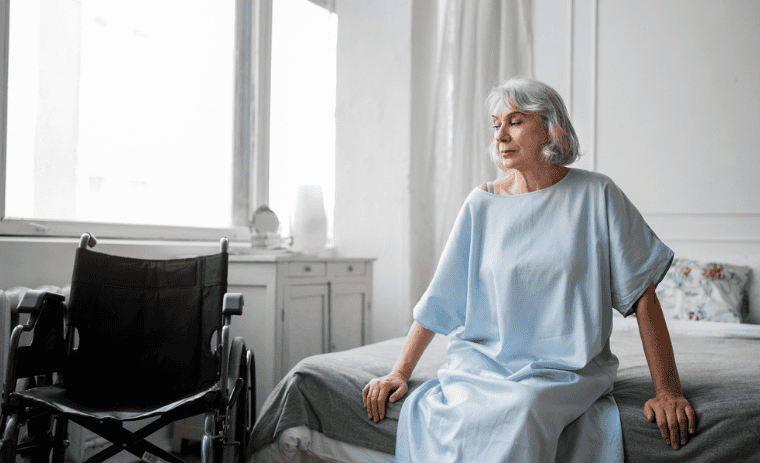 Senior woman sitting on bed next to wheelchair in bright bedroom - bed alarms and fall prevention for elderly home care