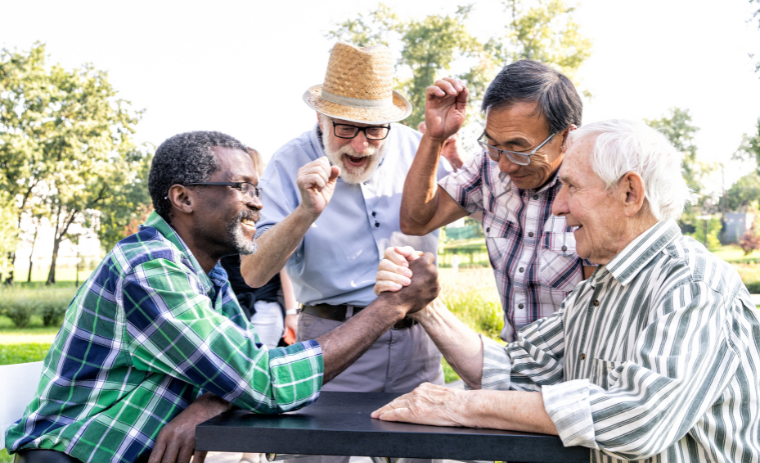 Group of smiling seniors enjoying fun summer activities for seniors outdoors, arm-wrestling and cheering together at a park.